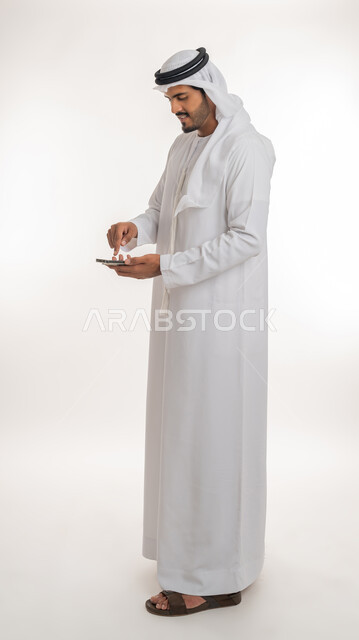 The development of technology and modern techniques, browsing social networking sites via the Internet, a portrait of an Emirati, Arab, Gulf man wearing a kandora and ghutra, using a mobile phone, making video calls via mobile, full-length body photo, white background