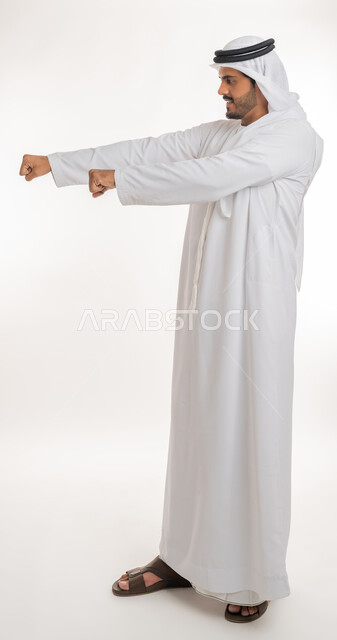 ممارسة رياضة الدفاع عن النفس، تعزيز صحة العظام والمفاصل، إتقان فنون الملاكمة، زيادة قدرة العضلات على التحمل، بورتريه من الجانب لرجل عربي خليجي اماراتي يرتدي الكندورة والغترة يرفع قبضات يديه بإيماءات القوة، صورة الجسم بالطول الكامل, خلفية بيضاء