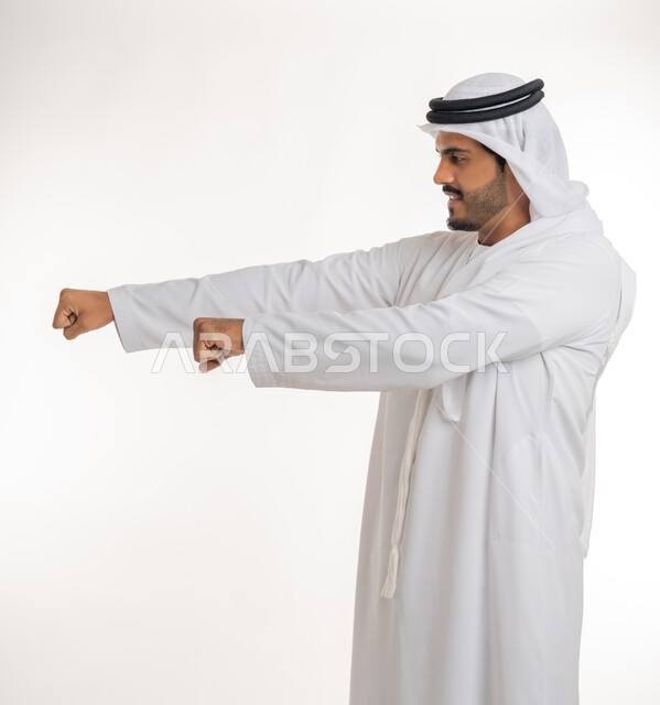 تعزيز صحة العظام والمفاصل، إتقان فنون الملاكمة، ممارسة رياضة الدفاع عن النفس، زيادة قدرة العضلات على التحمل، بورتريه من الجانب لرجل عربي خليجي اماراتي يرتدي الكندورة والغترة يرفع قبضات يديه بإيماءات القوة، خلفية بيضاء