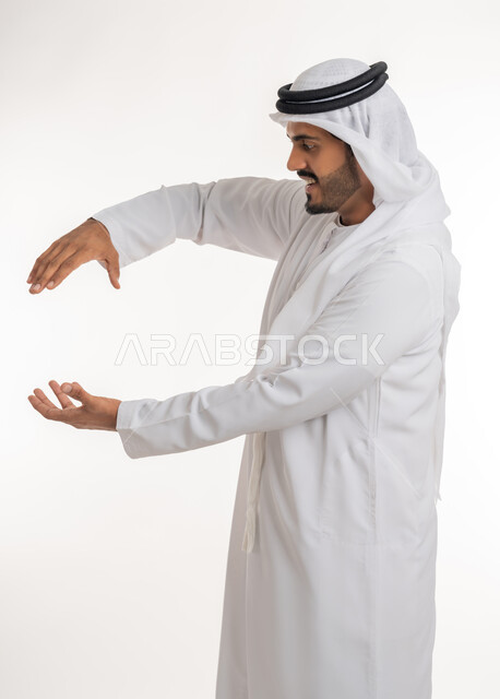 مفهوم الاناقة و الاهتمام بالمظهر الخارجي, إحتواء وإمساك عنصر ما،  بورتريه مقرب لرجل عربي خليجي اماراتي يرتدي الكندورة و العمامة يفتح يديه لعرض عناصر هولوجرامية ثلاثية الأبعاد، خلفية بيضاء