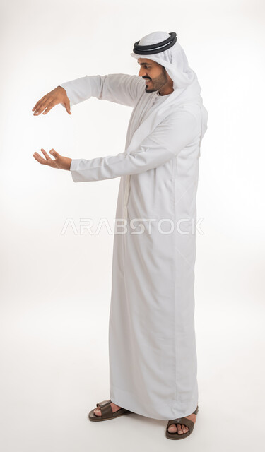 إحتواء وإمساك عنصر ما، مفهوم الاناقة و الاهتمام بالمظهر الخارجي, بورتريه مقرب لرجل عربي خليجي اماراتي يرتدي الكندورة و العمامة يفتح يديه لعرض عناصر هولوجرامية ثلاثية الأبعاد، صورة الجسم بالطول الكامل, خلفية بيضاء