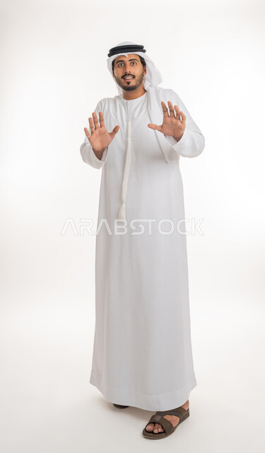 A request to stop and refrain from approaching, to protect oneself from external harm, a portrait of a young Arab Gulf Emirati teenager wearing a kandura and a ghutra, looking at the camera and raising his hands with an expression of fear and distance, full-length body portrait, white background.