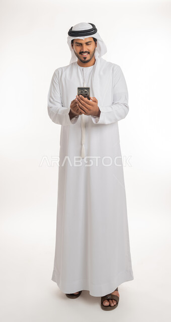تطور التكنولوجيا والتقنيات الحديثة، تصفح مواقع التواصل الاجتماعي عبر الانترنت، بورتريه لرجل اماراتي عربي خليجي يرتدي الكندورة والغترة  يقوم باستخدام الهاتف المحمول، إجراء مكالمات فيديو عبر الجوال، صورة الجسم بالطول الكامل، خلفية بيضاء