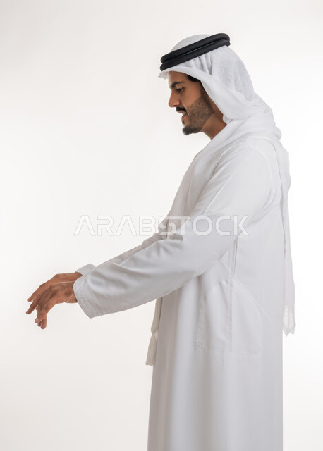الوقوف باستقامه والنظر الى الكاميرا بتعابير الثقه بالنفس، مفهوم الاناقه والاهتمام بالمظهر الخارجي، بورتريه لرجل عربي خليجي اماراتي يرتدي الكندوره والغتره البيضاء الاماراتيه يقف بايماءات الرجولة، خلفية بيضاء