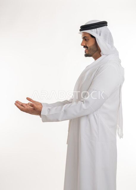 مفهوم الطلب والحاجة إلى مساعدة, تقديم وعرض شيء، بورتريه من الجانب لرجل عربي خليجي امارتي مبتسم يرتدي الكندورة و الغترة  يفتح الكفين بإيماءات محاولة حمل عنصر ما، خلفية بيضاء