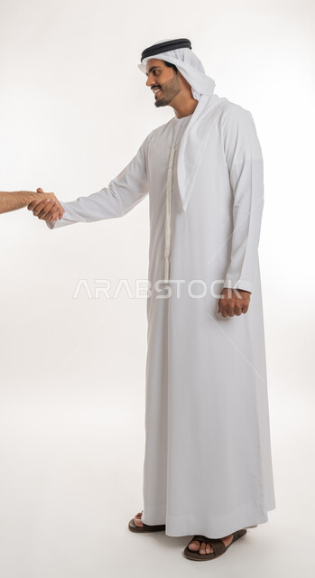 Greeting and peace, reception and welcome concept, portrait of an Arab Gulf Emirati man wearing a white kandura and ghutra looking at extending his hand with shaking gestures, acquaintance, friendship and forming relationships, full body, white background