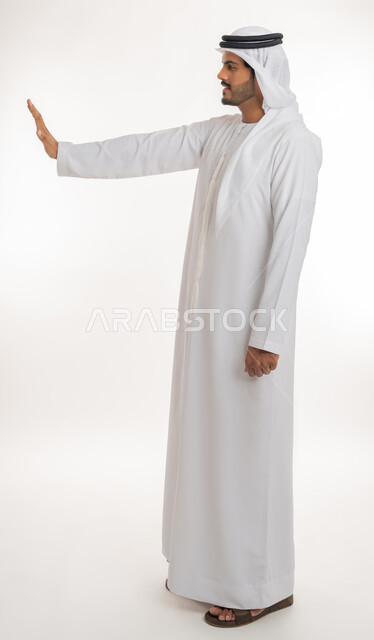 مفهوم الرفض والامتناع والانزعاج، النهي عن فعل أمر ما، بورتريه من الجانب لرجل عربي خليجي إماراتي يرتدي الكندورة والغترة يرفع يده بإيماءات التوقف وعدم الاقتراب، صورة الجسم بالطول الكامل، خلفية بيضاء