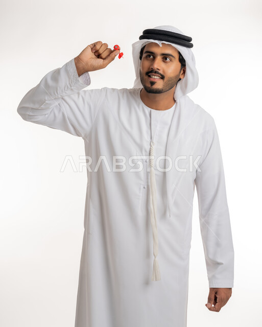 The concept of aiming and focusing, accuracy in determining the target, hobbies and sports skills, entertainment and fun and challenge in throwing arrows, portrait of an Arab Gulf Emirati man wearing a white kandura holding a red magnetic arrow, white background