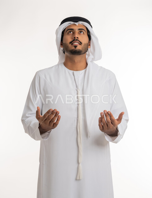 الدعاء ورفع اليدين إلى السماء، العبادة وترديد الأدعية بإيماءات الخضوع، بورتريه مقرب لرجل عربي خليجي إماراتي يرتدي الكندورة والغترة يرفع يديه وينظر للأعلى بتعابير التضرع لله تعالى، خلفية بيضاء