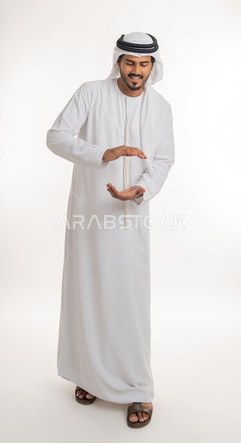 إحتواء وإمساك عنصر ما، مفهوم الاناقة و الاهتمام بالمظهر الخارجي, بورتريه لشاب عربي خليجي اماراتي يرتدي الكندورة و الغترة يفتح يديه لعرض عناصر هولوجرامية ثلاثية الأبعاد، صورة الجسم بالطول الكامل, خلفية بيضاء