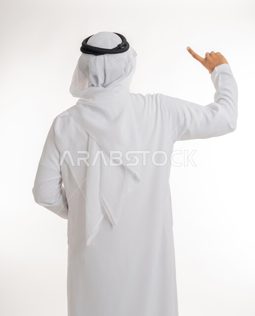 الإشارة إلى عنصر ما، بورتريه مقرب من الخلف لرجل عربي خليجي اماراتي يرتدي الكندورة والغترة البيضاء يرفع يده للاعلى يشير بأصبع السبابة الى هدف محدد، خلفية بيضاء