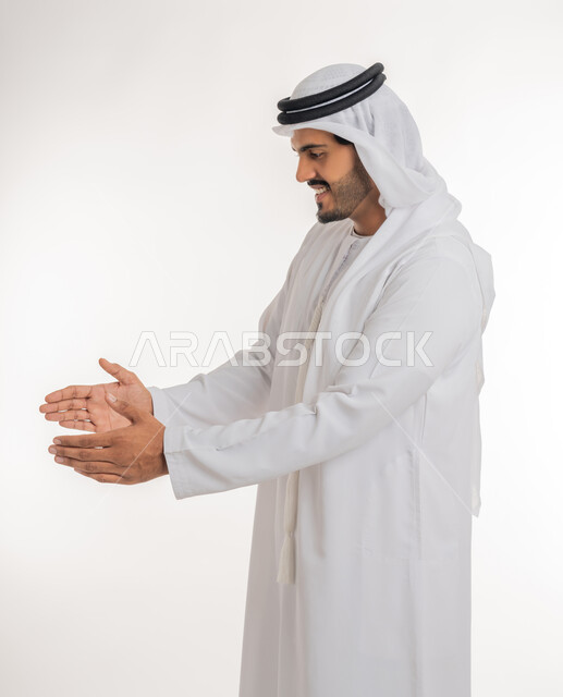 مفهوم الاناقة و الاهتمام بالمظهر الخارجي, إحتواء وإمساك عنصر ما،  بورتريه مقرب لرجل عربي خليجي اماراتي يرتدي الكندورة و العمامة يفتح يديه لعرض عناصر هولوجرامية ثلاثية الأبعاد، خلفية بيضاء