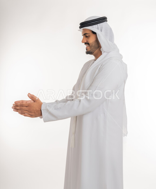 إحتواء وإمساك عنصر ما، مفهوم الاناقة و الاهتمام بالمظهر الخارجي, بورتريه من الجانب لرجل عربي خليجي اماراتي يرتدي الكندورة و الغترة يفتح يديه لعرض عناصر هولوجرامية ثلاثية الأبعاد، خلفية بيضاء