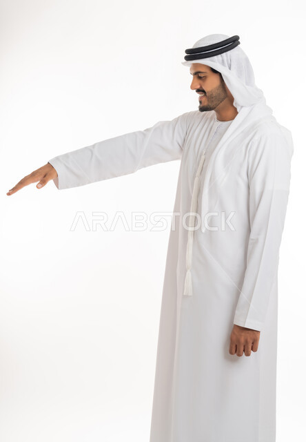 تحديد طول وإرتفاع شيء ما، تعابير واشارات يدين تدل على قياس المسافة، بورتريه من الجانب لرجل عربي خليجي امارتي يرتدي الكندورة والغترة الامارتية يمد راحة كفه نحو الأسفل بايماءات وجود عنصر معين، خلفية بيضاء