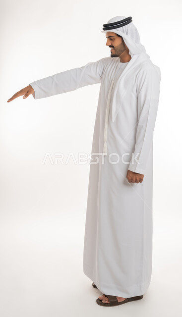 تعابير واشارات يدين تدل على قياس المسافة، تحديد طول وإرتفاع شيء ما، بورتريه من الجانب لرجل عربي خليجي امارتي يرتدي الكندورة والغترة الامارتية يمد راحة كفه نحو الأسفل بايماءات وجود عنصر معين، صورة الجسم بالطول الكامل, خلفية بيضاء
