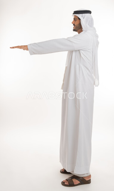 تحديد طول وإرتفاع شيء ما، تعابير واشارات يدين تدل على قياس المسافة،  بورتريه من الجانب لرجل عربي خليجي امارتي يرتدي الكندورة والغترة الامارتية يمد راحة كفه نحو الأسفل بايماءات وجود عنصر معين، صورة الجسم بالطول الكامل, خلفية بيضاء