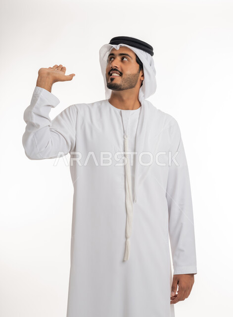 محاولة حمل عنصر ما عاليا، مفهوم الاناقة و الاهتمام بالمظهر الخارجي,  بورتريه مقرب لرجل عربي خليجي امارتي يرتدي الكندورة و الغترة ينظر  يرفع كفه للاعلى يحمل عناصر هولوجرامية ثلاثية الأبعاد، خلفية بيضاء