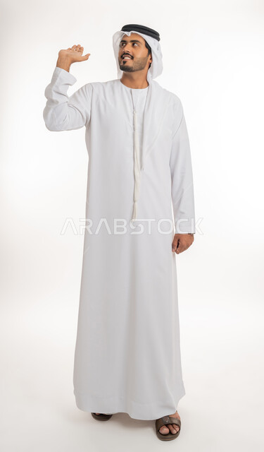 مفهوم الاناقة و الاهتمام بالمظهر الخارجي, محاولة حمل عنصر ما عاليا،  بورتريه لرجل عربي خليجي امارتي يرتدي الكندورة و الغترة ينظر  يرفع كفه للاعلى يحمل عناصر هولوجرامية ثلاثية الأبعاد، صورة الجسم بالطول الكامل, خلفية بيضاء