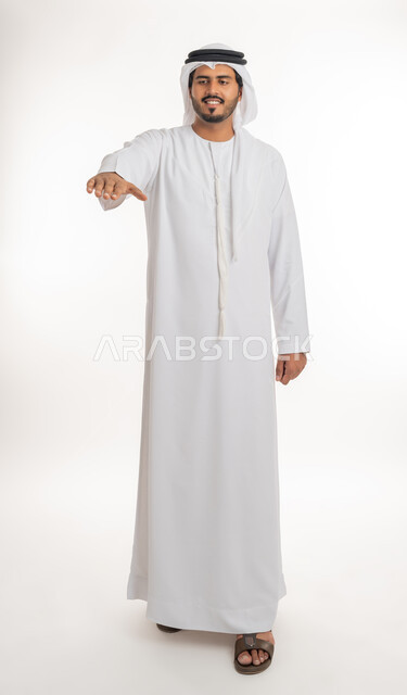 Determining the length and height of something, expressions and hand gestures that indicate measuring distance, a portrait of an Arab Gulf Emirati man wearing a kandora and an Emirati ghutra, extending his palm downward with gestures of the presence of a specific element, full-length body image, white background.