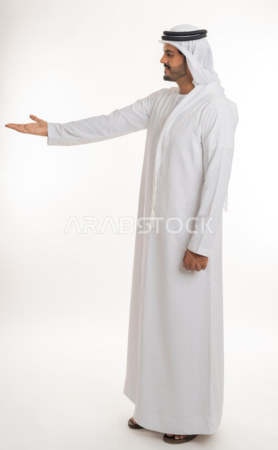 محاولة رفع عنصر ما، مفهوم الاناقة و الاهتمام بالمظهر الخارجي, بورتريه من الجانب لرجل عربي خليجي امارتي يرتدي الكندورة و الغترة ينظر  يرفع كفه للاعلى يحمل عناصر هولوجرامية ثلاثية الأبعاد، صورة الجسم بالطول الكامل, خلفية بيضاء