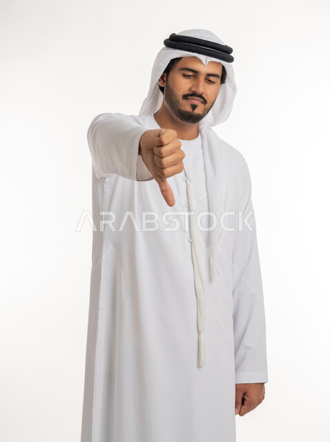حركات يدين تدل على الرفض وعدم القبول والفشل، بورتريه مقرب لرجل عربي خليجي امارتي يرتدي الكندورة و الغترة يشير بإصبعه الابهام الى الأسفل بإيماءات عدم الاعجاب والتقييم السلبي، الشعور بالانزعاج من شيء ما، خلفية بيضاء
