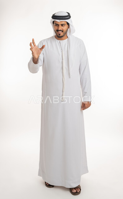 مفهوم الاناقة و الاهتمام بالمظهر الخارجي, إحتواء وإمساك عنصر ما،  بورتريه لرجل عربي خليجي امارتي يرتدي الكندورة و الغترة يمد يده للأمام يحاول التقاط عناصر هولوجرامية ثلاثية الأبعاد، صورة الجسم بالطول الكمال, خلفية بيضاء