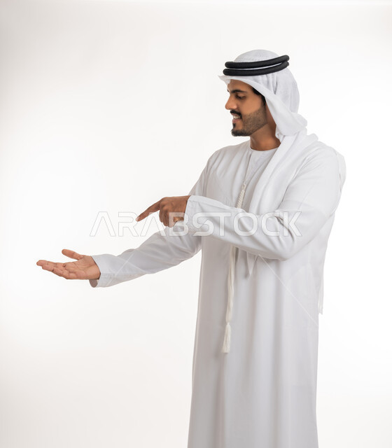 Gestures of carrying and lifting something, a portrait of an Arab Gulf Emirati man wearing a turban and a white Emirati kandura, opening one hand and pointing with his index finger towards the other hand, pointing towards a goal, finger movement and gestures, white background.