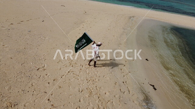 سائع عربي خليجي سعودي في رحلة إلى جزيرة سمر في منطقة عسير بالمملكة العربية السعودية، يحمل بيده علم السعودية، السياحة بالسعودية، الطبيعة الساحرة المطلة على جزيرة سمر الواقعة على ساحل منطقة عسير في وضع النهار