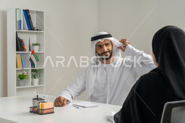 مفهوم التحاور وتبادل الأراء في خطط إنجاز الأعمال، إجراء مقابلة التعيين والتوظيف، صورة مقربة لمدير عربي خلجي إماراتي يرتدي الغترة والكندورة داخل المكتب يقوم بإجراء مقابلة عمل مع موظفة، مهن ووظائف إدارية مكتبية في الإمارات، المساواة بين المرأة والرجل