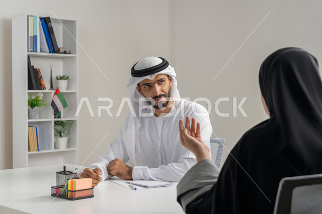 مهن ووظائف إدارية مكتبية في الإمارات، مفهوم التحاور وتبادل الأراء في خطط إنجاز الأعمال، إجراء مقابلة التعيين والتوظيف، صورة مقربة لمدير عربي خلجي إماراتي يرتدي الغترة والكندورة داخل المكتب يقوم بإجراء مقابلة عمل مع موظفة، المساواة بين المرأة والرجل