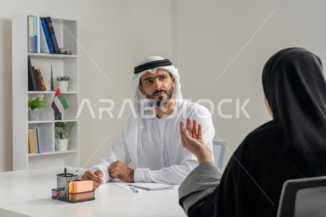 إجراء مقابلة التعيين والتوظيف، مفهوم التحاور وتبادل الأراء في خطط إنجاز الأعمال، صورة مقربة لمدير عربي خلجي إماراتي يرتدي الغترة والكندورة داخل المكتب يقوم بإجراء مقابلة عمل مع موظفة، مهن ووظائف إدارية مكتبية في الإمارات، المساواة بين المرأة والرجل