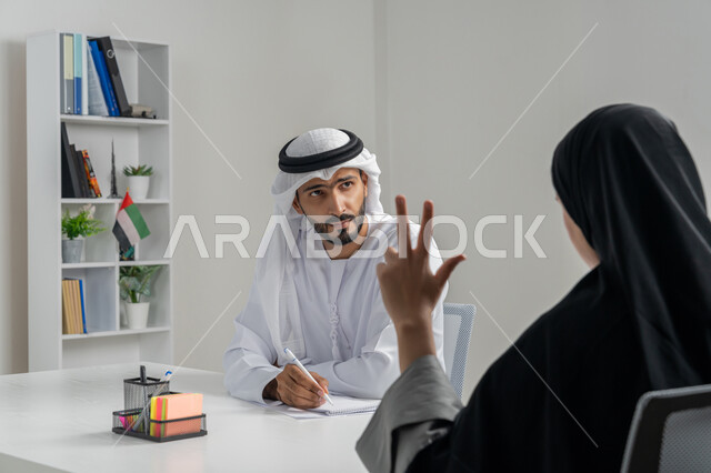مهن ووظائف إدارية مكتبية في الإمارات، مفهوم التحاور وتبادل الأراء في خطط إنجاز الأعمال، إجراء مقابلة التعيين والتوظيف، صورة مقربة لمدير عربي خلجي إماراتي يرتدي الغترة والكندورة داخل المكتب يقوم بإجراء مقابلة عمل مع موظفة، المساواة بين المرأة والرجل