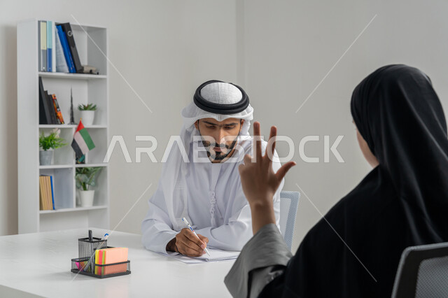 إجراء مقابلة التعيين والتوظيف، مفهوم التحاور وتبادل الأراء في خطط إنجاز الأعمال، صورة مقربة لمدير عربي خلجي إماراتي يرتدي الغترة والكندورة داخل المكتب يقوم بإجراء مقابلة عمل مع موظفة، مهن ووظائف إدارية مكتبية في الإمارات، المساواة بين المرأة والرجل