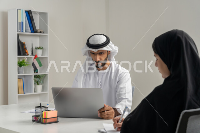 استخدام الحاسب المحمول ضمن الشركات، إجراء مقابلة التعيين والتوظيف، مفهوم التحاور وتبادل الأراء في خطط إنجاز الأعمال، صورة مقربة لمدير عربي خلجي إماراتي يرتدي الغترة والكندورة داخل المكتب يقوم بإجراء مقابلة عمل مع موظفة، مهن ووظائف إدارية مكتبية في الإمارات
