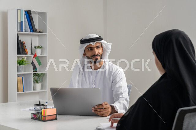 استخدام الحاسب المحمول ضمن الشركات، إجراء مقابلة التعيين والتوظيف، مفهوم التحاور وتبادل الأراء في خطط إنجاز الأعمال، صورة مقربة لمدير عربي خلجي إماراتي يرتدي الغترة والكندورة داخل المكتب يقوم بإجراء مقابلة عمل مع موظفة، مهن ووظائف إدارية مكتبية في الإمارات