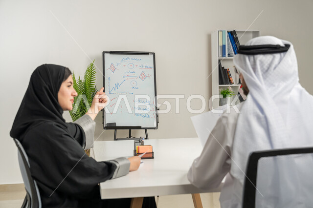 تبادل الأراء والافكار بين الموظفين، رجل عربي خليجي إماراتي يرتدي الكندورة والغترة يجلس بجانب زميلته في اجتماع داخل مقر الشركة، تعلم اساليب العمل والتطوير المهني، العمل الجماعي وروح الفريق الواحد، وضع الخطط الاستراتيجية لتحقيق الأهداف، كتابة وتدوين الملاحظات