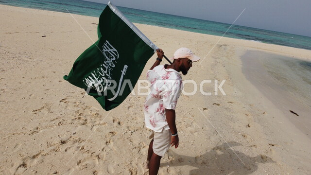 سائع عربي خليجي سعودي في رحلة إلى جزيرة سمر في منطقة عسير بالمملكة العربية السعودية، يحمل بيده علم السعودية، السياحة بالسعودية، الطبيعة الساحرة المطلة على جزيرة سمر الواقعة على ساحل منطقة عسير في وضع النهار