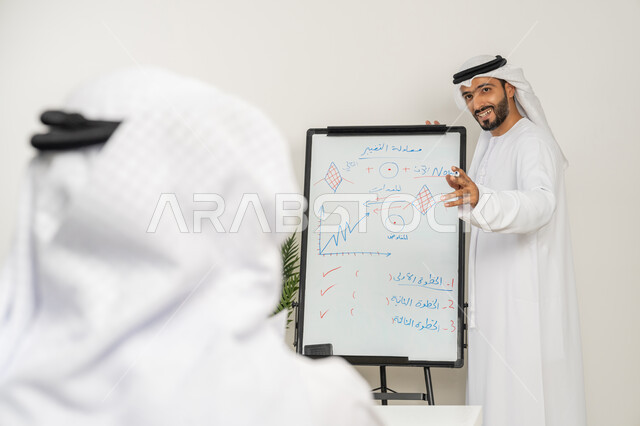 Ensuring a clear understanding of the business using the whiteboard, developing strategic plans to achieve goals, two Emirati Gulf Arab men wearing kandura and ghutra holding a meeting inside the company headquarters, learning work methods and professional development, teamwork and team spirit, exchanging opinions between employees