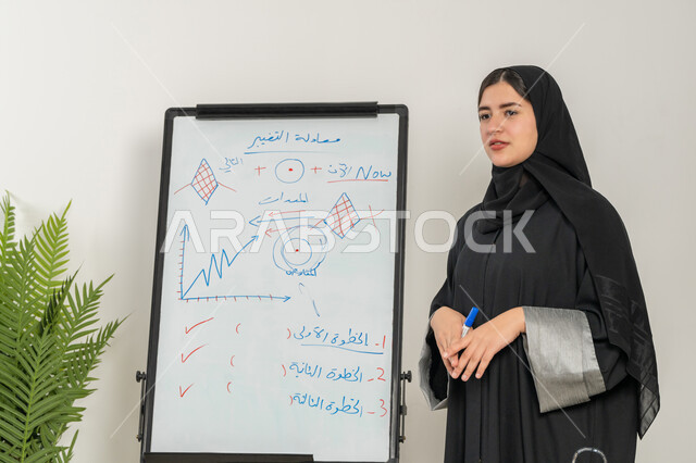 مشاركة النساء في المجالات العملية, توضيح الخطوات لتحقيق الأهداف، مدربة سعودية تستخدم السبورة لسهولة الشرح، صورة مقربة لإمرأة عربية خليجية إماراتية ترتدي الحجاب والعباءة تقوم بورشات عمل تدريبية مهنية داخل مقر الشركة, تطوير استراتيجيات شركات الإمارات العربية المتحدة