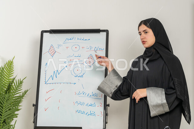 توضيح الخطوات لتحقيق الأهداف، مدربة سعودية تستخدم السبورة لسهولة الشرح، صورة مقربة لإمرأة عربية خليجية إماراتية ترتدي الحجاب والعباءة تقوم بورشات عمل تدريبية مهنية داخل مقر الشركة, تطوير استراتيجيات شركات الإمارات العربية المتحدة، تعلم اساليب العمل والتطوير المهني