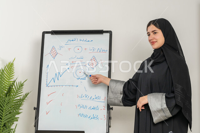 توضيح الخطوات لتحقيق الأهداف، مدربة سعودية تستخدم السبورة لسهولة الشرح، صورة مقربة لإمرأة عربية خليجية إماراتية ترتدي الحجاب والعباءة تقوم بورشات عمل تدريبية مهنية داخل مقر الشركة, تطوير استراتيجيات شركات الإمارات العربية المتحدة، تعلم اساليب العمل والتطوير المهني