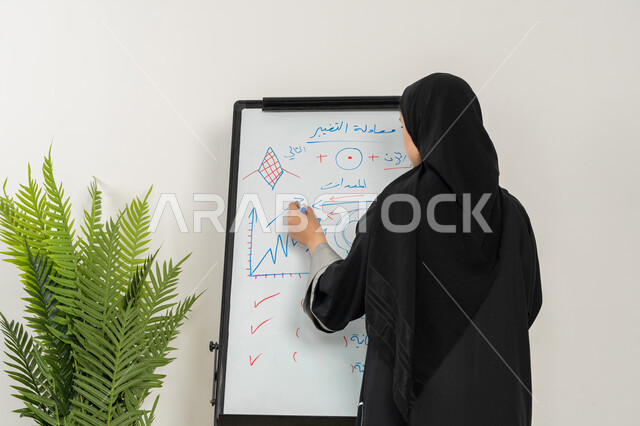 مدربة سعودية تستخدم السبورة لسهولة الشرح، ورشات عمل تدريبية مهنية، صورة مقربة لإمرأة عربية خليجية إماراتية ترتدي الحجاب والعباءة تقوم بتوضيح الخطوات لتحقيق الأهداف داخل مقر الشركة, تطوير استراتيجيات شركات الإمارات العربية المتحدة، تعلم اساليب العمل والتطوير المهني