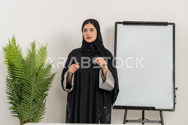 تطوير استراتيجيات شركات الإمارات العربية المتحدة، تعلم اساليب العمل والتطوير المهني، ورشات عمل تدريبية مهنية، مدربة سعودية تستخدم السبورة لسهولة الشرح، صورة مقربة لإمرأة عربية خليجية إماراتية ترتدي الحجاب والعباءة تقوم بتوضيح الخطوات لتحقيق الأهداف داخل مقر الشركة