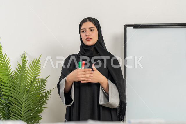 ورشات عمل تدريبية مهنية، مدربة سعودية تستخدم السبورة لسهولة الشرح، صورة مقربة لإمرأة عربية خليجية إماراتية ترتدي الحجاب والعباءة تقوم بتوضيح الخطوات لتحقيق الأهداف داخل مقر الشركة, تطوير استراتيجيات شركات الإمارات العربية المتحدة، تعلم اساليب العمل والتطوير المهني