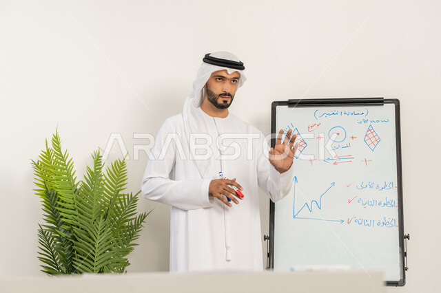 تطوير استراتيجيات شركات الإمارات العربية المتحدة، تعلم اساليب العمل والتطوير المهني، ورشات عمل تدريبية مهنية، استخدام السبورة لسهولة الشرح، صورة مقربة لرجل عربي خليجي إماراتي يرتدي الغترة والكندورة يقوم بتوضيح الخطوات لتحقيق الأهداف داخل مقر الشركة