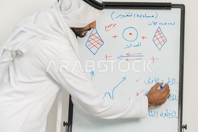 تعلم اساليب العمل والتطوير المهني، ورشات عمل تدريبية مهنية، استخدام السبورة لسهولة الشرح، صورة مقربة لرجل عربي خليجي إماراتي يرتدي الغترة والكندورة يقوم بتوضيح الخطوات لتحقيق الأهداف داخل مقر الشركة, تطوير استراتيجيات شركات الإمارات العربية المتحدة