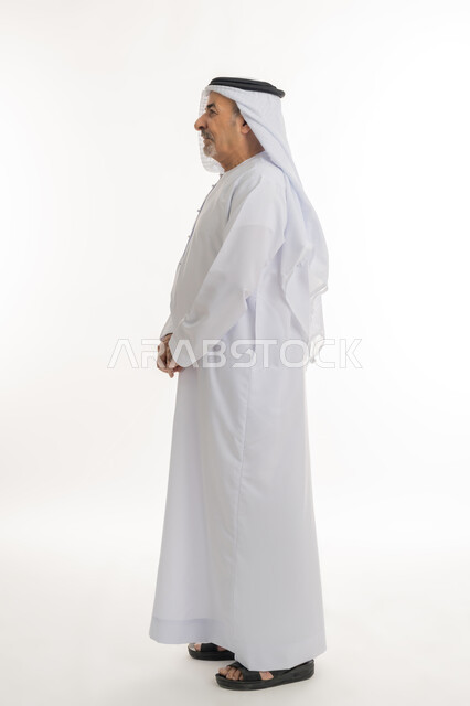 إيماءات الجدية والحزم، التقدم بالعمر والهيبة والوقار، بورتريه من الجانب لرجل كبير بالسن عربي خليجي إماراتي يرتدي الكندورة ينظر الى شيء ما, الاهتمام بالأناقة والمظهر الخارجي، الوقوف باستقامة وثبات، مفهوم الرجولة العربية, الطول الكامل للجسم، خلفية بيضاء