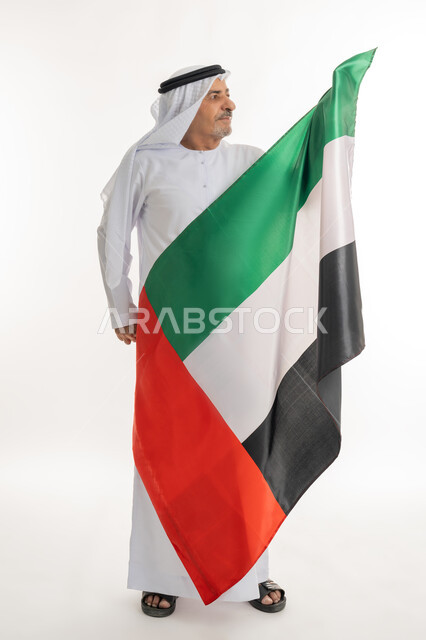 الإحتفال باليوم الوطني الإماراتي، يوم استقلال الإمارات، بورتريه لرجل عربي خليجي إماراتي يرتدي الكندورة والغترة البيضاء يحمل علم الإمارات العربية المتحدة وينظر إليه، الاعتزاز والفخر بإنجازات البلاد، هوية اليوم الوطني الاماراتي 51، الطول الكامل للجسم، خلفية بيضاء