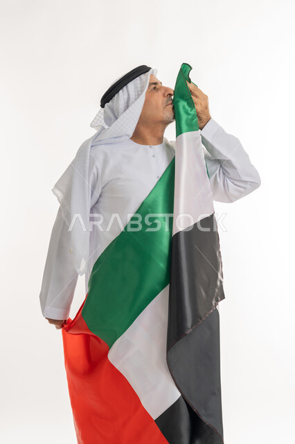 الاعتزاز والفخر بإنجازات البلاد، هوية اليوم الوطني الاماراتي 51، الإحتفال باليوم الوطني الإماراتي، يوم استقلال الإمارات، الحب والانتماء للوطن، بورتريه لرجل عربي خليجي إماراتي يرتدي الكندورة والغترة البيضاء يقوم بتقبيل علم الإمارات العربية المتحدة، خلفية بيضاء