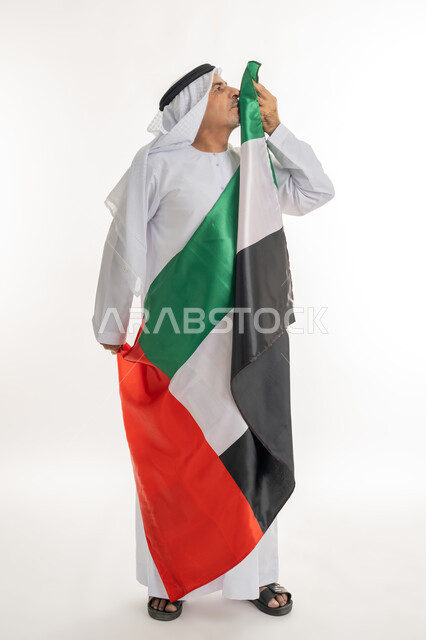 هوية اليوم الوطني الاماراتي 51، الإحتفال باليوم الوطني الإماراتي، يوم استقلال الإمارات، بورتريه لرجل عربي خليجي إماراتي يرتدي الكندورة والغترة البيضاء يقوم بتقبيل علم الإمارات العربية المتحدة، الاعتزاز والفخر بإنجازات البلاد، الطول الكامل للجسم، خلفية بيضاء